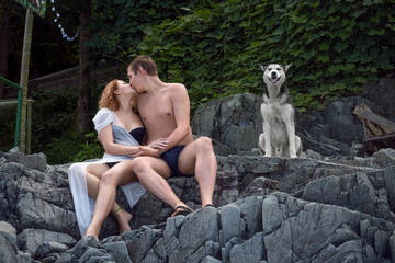 A young couple is sitting on the stone steps of a cliff overgrown with vines, and kissing, with a faithful dog sitting next to them