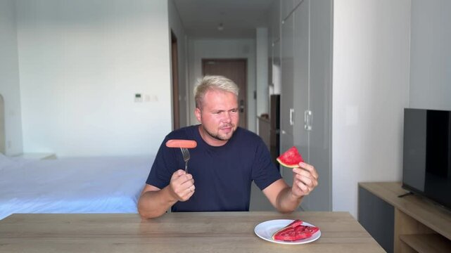 A handsome man, a stylish blond man in a blue t-shirt is sitting at a table in his flat with a bright modern interior and decides what to eat, watermelon or sausage, he wants sausage but chooses to ea