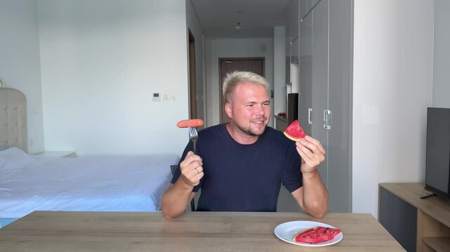 A handsome man, a stylish blond man in a blue t-shirt is sitting at a table in his flat with a bright modern interior and decides what to eat, watermelon or sausage, he wants sausage but chooses to ea