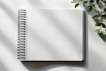 Blank spiral-bound notebook lying open on a white surface with natural light and green leafy plant casting soft shadows