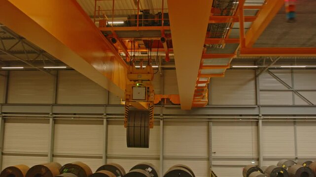A large overhead bridge crane transports a heavy steel coil through an automated warehouse