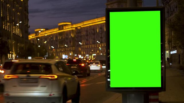 Busy city street at night with moving cars and bright lights, featuring large green screen billboard for advertising. Concept of modern media and urban energy