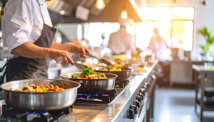A chef in a professional kitchen cooking food in pans on a stove line with sunlight streaming in.
