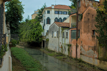 Fototapeta premium This serene scene captures a quiet canal flowing through the historic city of Padua, Italy. Lush greenery drapes over the banks, contrasting with the aged buildings lining the waterway.