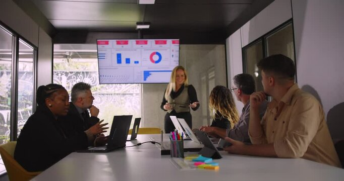 Blonde professional smiling during presentation while team listens and digital charts display key metrics in modern office setting