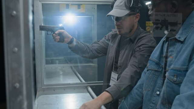 Backlit side medium of professional Caucasian male shooting range instructor conducting first gun shooting lesson for African American male customer, explaining basics of firearm handling and aiming