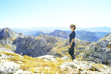 Fototapeta premium Hiking in the most beautiful places of Montenegro: a young smiling woman stands on the trail under the peak of Bobotov Kuk. Eco-tourism in Zabljak: a female tourist in the Durmitor National Park