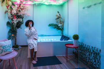 Young woman wearing bathrobe drinking champagne and relaxing in a modern beauty spa with chromotherapy lights