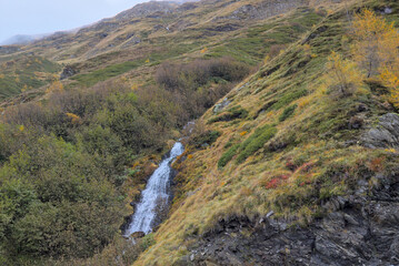 A vibrant waterfall tumbles down a steep, green hillside, accented by autumn foliage in gold and...