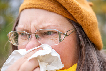 Sick mature woman with tissue, blowing nose, suffering from seasonal allergy, outdoor portrait,...