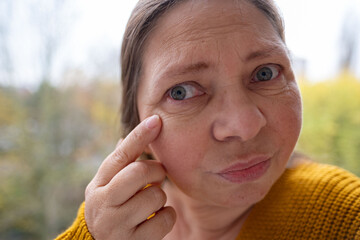 mature female face woman 50-55 years old, looks examines wrinkles around eyes, skin age-related changes, care anti-aging procedures