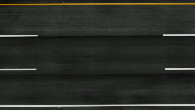 Aerial drone view of a straight asphalt road with clear white lane markings, showcasing smooth, well-maintained infrastructure surrounded by dark pavement textures from above.
