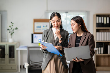 Two women in business attire are looking at a blue folder and a laptop