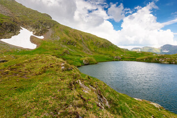 alpine lake in fagaras mountains. grassy slopes with stones and snow under cloudy sky in summer....