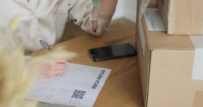 Close-up video of a female customer writing on paper and filling out a form, wanting to return a product. A young woman writes a return order and sits at a table with a laptop at home.
