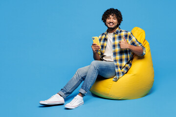 Full body young Indian man he wears yellow checkered shirt casual clothes sit in bag chair hold in...