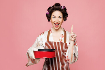 Young housewife housekeeper chef cook baker woman wears brown apron hold red metal baking form for pie, baking dish point finger up isolated on pink background studio portrait. Cooking food concept.