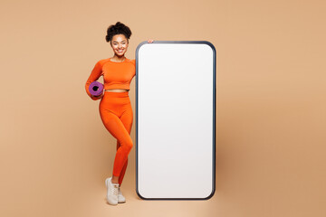 Full body young fitness trainer instructor woman in orange clothes spend time in gym big huge blank screen mobile cell phone hold mat isolated on plain beige background Workout sport fit abs concept