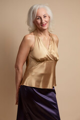 Portrait of Smiling Senior Caucasian Blond Woman Posing in Casual Brown Blouse And Vilet Skirt In Studio Over Beige Background With Behind Crossed Hands