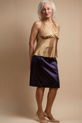 Full Length Portrait of Senior Caucasian Blond Woman Posing in Casual Brown Blouse And Vilet Skirt In Studio Over Beige Background With Behind Crossed Hands and Calm Facial Expression.