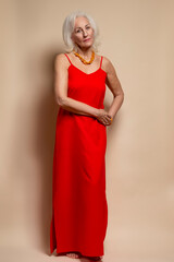Ageing Concepts. One Winsome Senior Caucasian Blond Woman Posing in Red Dress In Studio Over Beige Background With Positive Facial Expression.