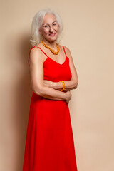 Ageing Concepts. Smiling Happy Winsome Senior Caucasian Blond Woman Posing in Red Dress In Studio Over Beige Background With Positive Facial Expression.