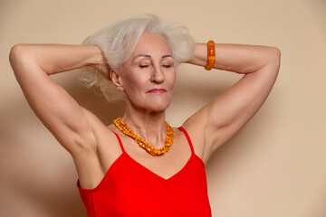 Ageing Concepts. Smiling Happy Winsome Senior Caucasian Blond Woman Posing in Red Dress With Lifted Hands And Eyes Closed In Studio Over Beige Background With Positive Facial Expression.