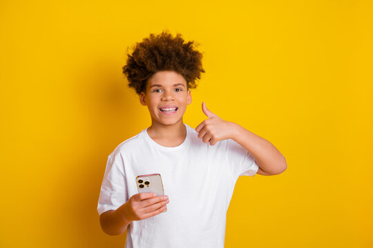 Cheerful boy holding smartphone and showing thumbs up on vibrant yellow background, expressing happiness and connection