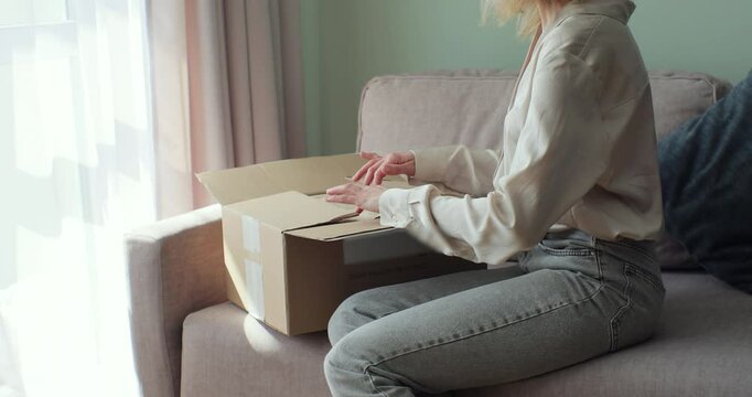 Young woman buyer opening parcel box at home. Female customer shopper receiving online shop purchase buying clothes unpacking delivery package.