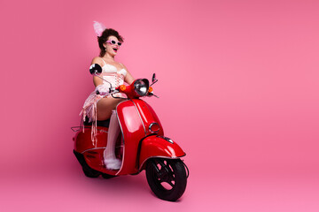 A young woman in a corset dress enjoys a fun ride on a vintage red scooter against a vibrant pink...