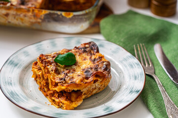 A piece of lasagna according to a traditional Italian recipe on a plate on a light background and in a baking dish. Close-up.