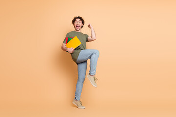 Excited young man jumping cheerfully while holding colorful folders against bright beige background in casual outfit