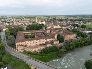 drone hovers above the crumbling stone walls of the 13th-century Pontevico Castle beside the slow meander of the Oglio River, with the town rooftops stretching eastward under a soft overcast sky.