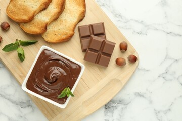 Tasty chocolate spread, pieces, toasts, mint and hazelnuts on white marble table, top view