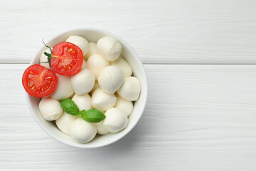 Tasty mozzarella cheese balls in bowl, basil and tomato on white wooden table, top view. Space for...