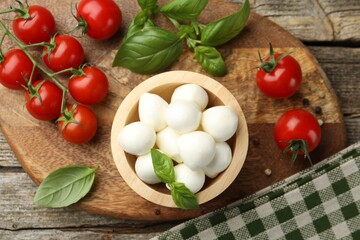 Tasty mozzarella cheese balls in bowl, basil, peppercorns and tomatoes on wooden table, top view