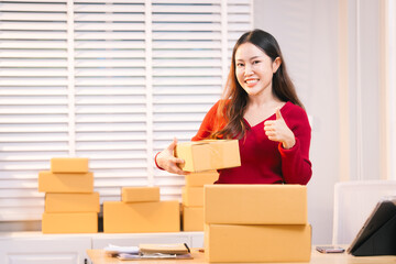 A smiling woman, who appears to be a middle-aged Asian individual, stacks cardboard boxes on a desk...