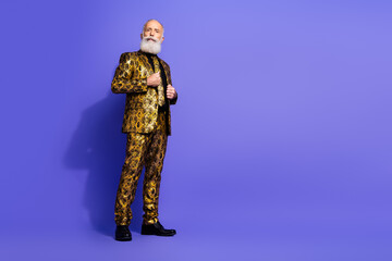 Elderly man in extravagant golden suit posing against purple background expressing confidence and a sense of flamboyant, stylish charm