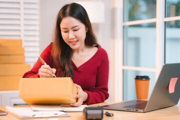 A smiling woman, who appears to be a middle-aged Asian individual, stacks cardboard boxes on a desk...