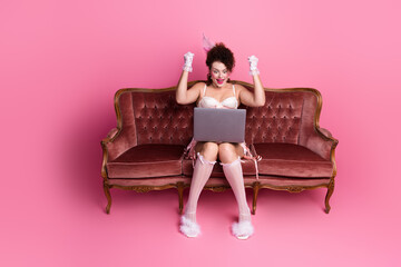 Obraz premium A woman dressed in an elegant vintage costume joyfully reacts to her laptop. The pink background and plush sofa add a touch of glamour
