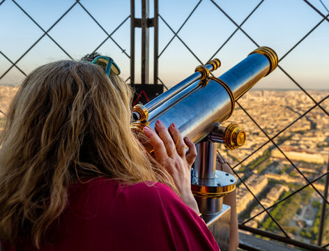 Blonde woman wearing a red t-shirt looking through a classic vintage metal telescope from the Eiffel Tower observation deck at the city of Paris from an aerial view at sunset during golden hour.