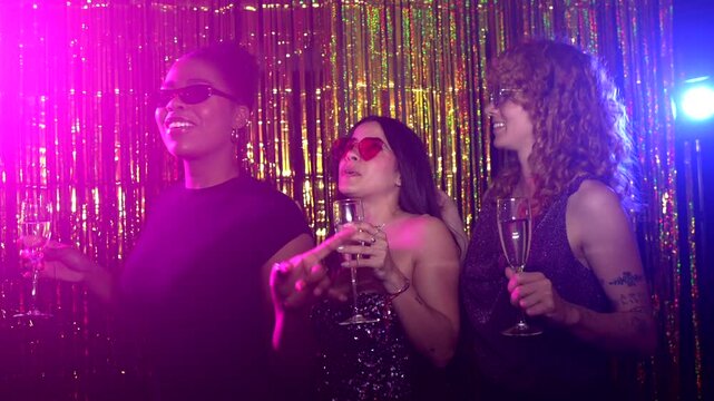 Young women wearing sunglasses and holding champagne glasses are having fun in a photo booth with a gold tinsel curtain