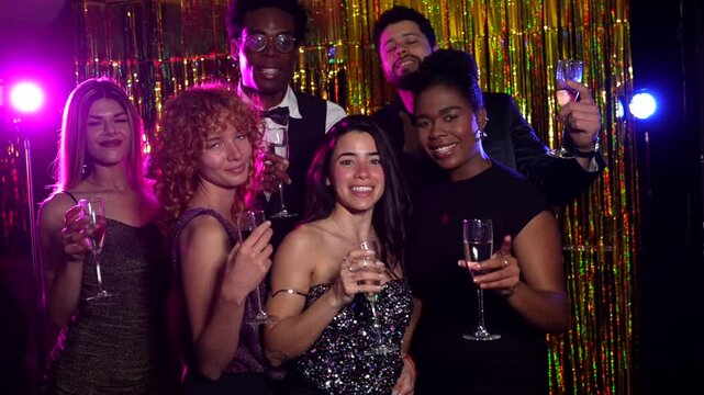 Cheerful friends are celebrating, raising glasses of champagne, posing together in a photo booth with shiny golden background