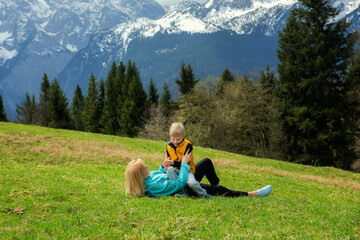 Naklejka premium Grandmother lying on green grass, playing with her happy grandson in yellow vest, surrounded by alpine mountains, trees, and nature. Joyful spring family moment outdoors.