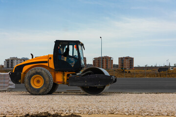 Yellow road roller compactor working on a gravel surface at a road construction site with buildings in the background.