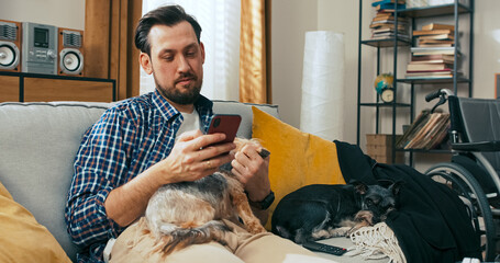 A middle-aged person with a disability uses a phone while sitting on the couch with several dogs, enjoying a moment of quiet companionship and focus in a home environment.