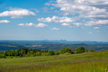 Fototapeta premium Panoramic view of green fields and forested hills near Jested, with the sharp peak of Ralsko mountain visible in the distance under a blue sky with scattered clouds.