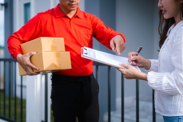 A uniformed delivery driver hands a package and clipboard to a woman outside her house, who is in the process of signing for the delivery. The open trunk of vehicle suggests more deliveries await.