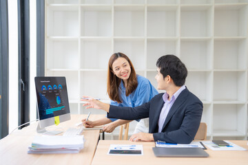 Two asian businesspeople discussing marketing strategy pointing at computer screen showing charts and graphs, working together in modern office using computer, tablet and paperwork