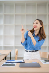 Asian businesswoman standing at her desk pointing up with both index fingers while smiling, working...